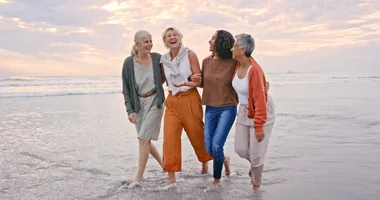 Four women laughing and walking arm-in-arm along a beach at sunset.