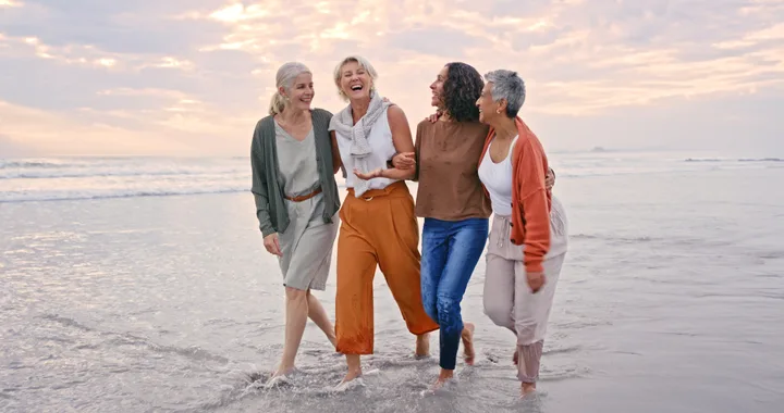 Four women laughing and walking arm-in-arm along a beach at sunset.
