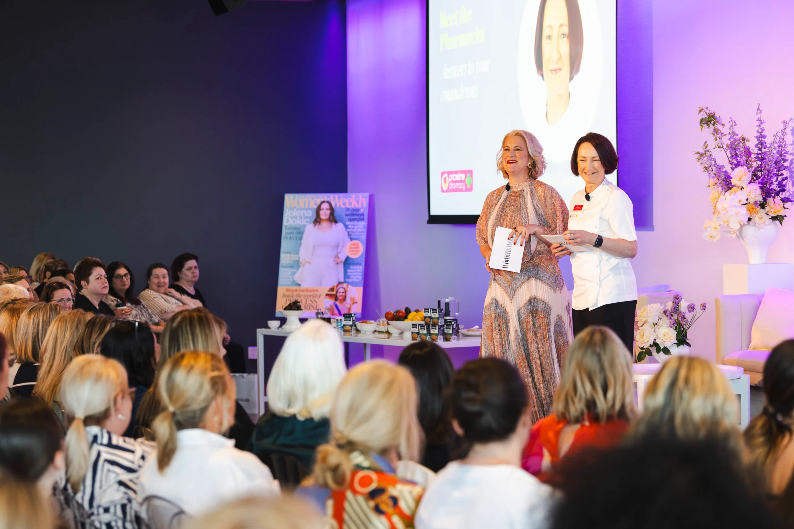 Two women present on stage at a wellness event, with an audience and a Woman's Weekly magazine cover in the background. Tickets for 2025 Health Summit available now