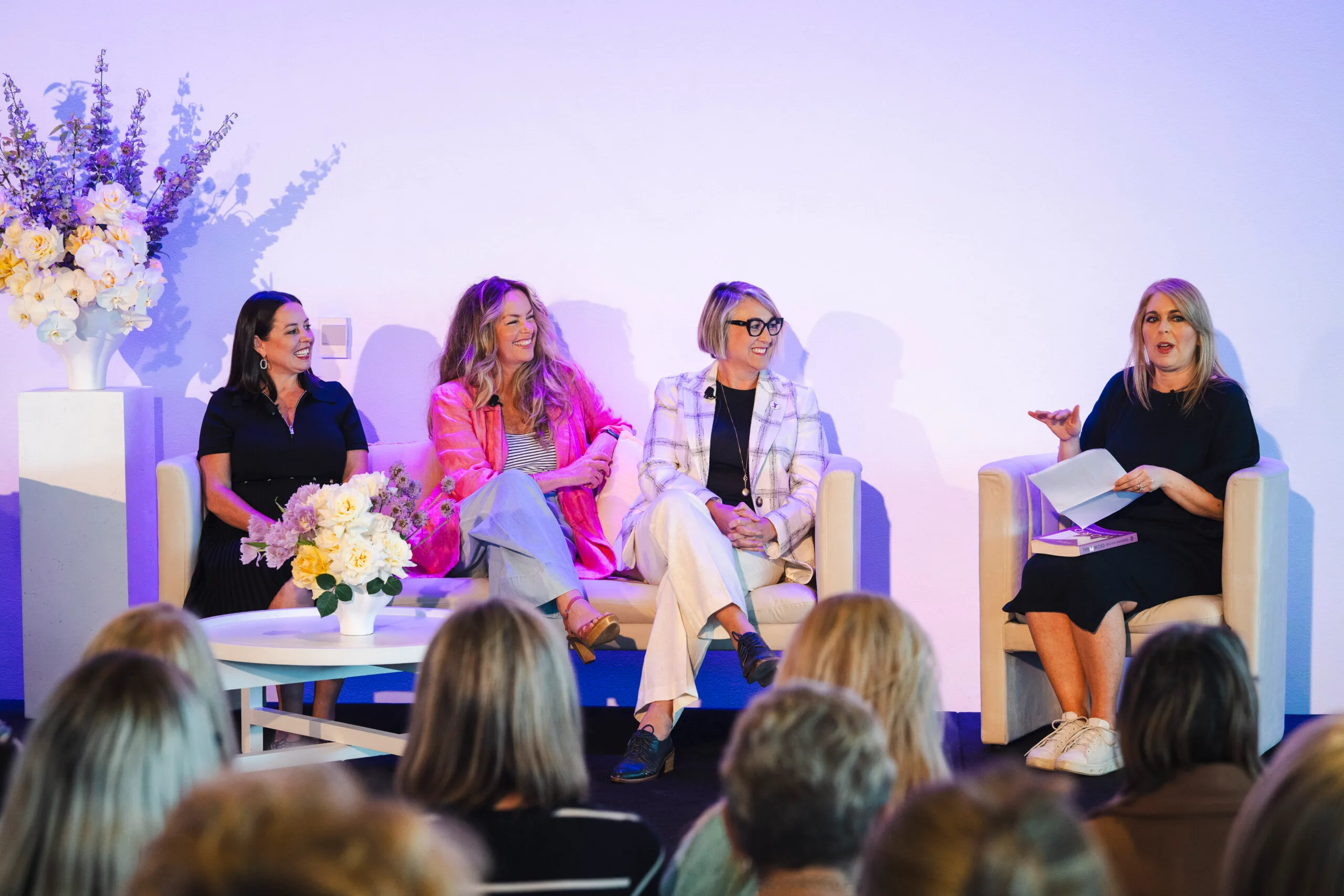 Four women seated on stage with flowers and speaking to an audience.