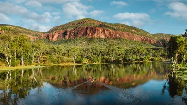 A kayak on a calm lake with lush green trees and a rocky hill under a partly cloudy sky in the background.