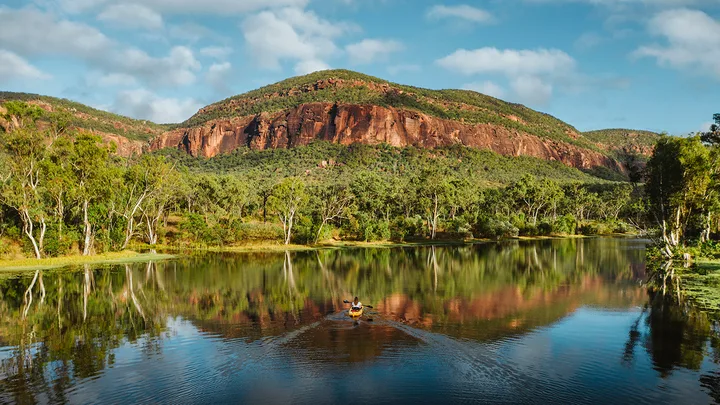 A kayak on a calm lake with lush green trees and a rocky hill under a partly cloudy sky in the background.