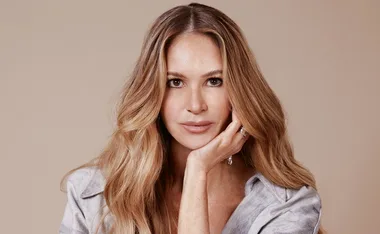A woman with long, wavy blonde hair and a thoughtful expression, resting her chin on her hand in a neutral background.