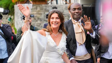 A smiling bride and groom wave at their wedding, surrounded by photographers.