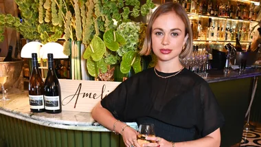 Woman in a black dress holds a wine glass at a bar with bottles labeled "Amelia" and lush green plants in the background.