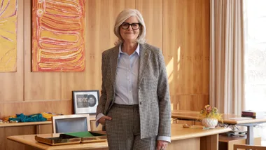 A person with gray hair and glasses stands smiling in an office, wearing a checked suit.