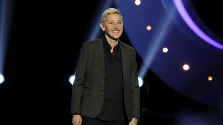 A person with short blonde hair in a dark suit smiles on stage with bright lights in the background.