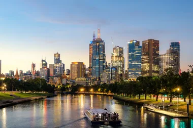 Skyline of Melbourne at dusk with a riverboat in the foreground and illuminated skyscrapers in the background.