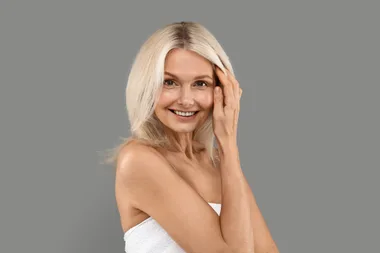 Smiling woman with blonde hair in a white towel, posing with hand on head against a gray background.