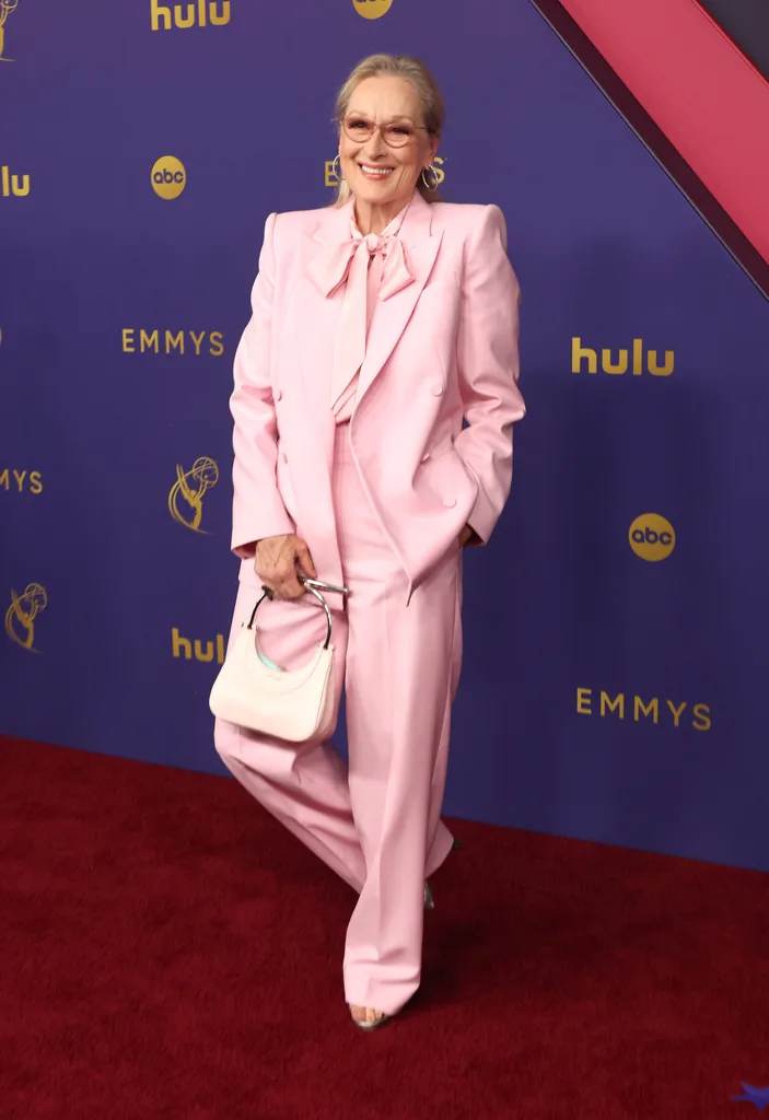 Person in a pink suit on Emmys red carpet, holding a white purse, smiling against a blue background with logos.