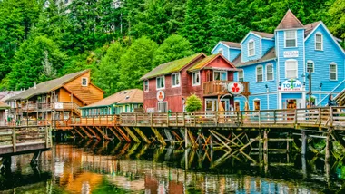 Colorful wooden buildings on a boardwalk alongside a forest and reflective water in Ketchikan, Alaska.