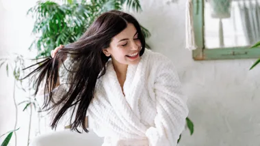 Woman in a white robe smiling and playing with her long, dark hair in a bright setting with green plants.