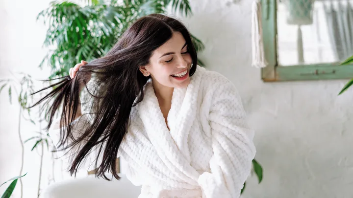 Woman in a white robe smiling and playing with her long, dark hair in a bright setting with green plants.