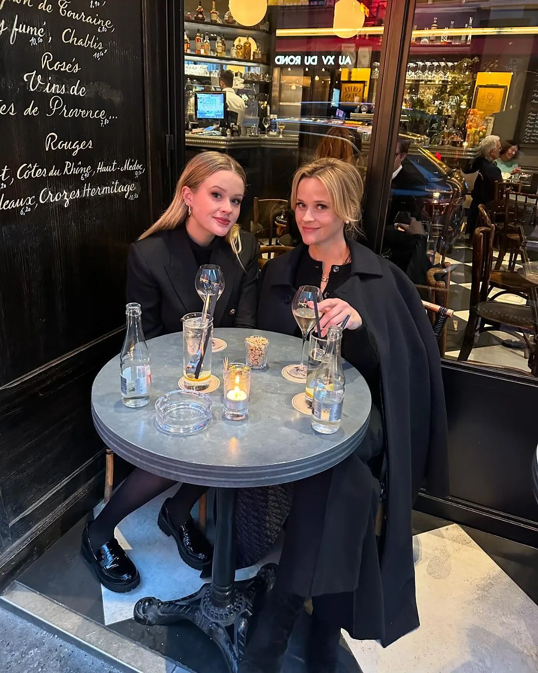 Two women in black outfits sit at a caf&eacute; table with drinks and candles on a dimly lit evening.