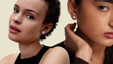Two women wearing pearl earrings, one with curly hair and a black top, the other with straight hair and a gold ring.