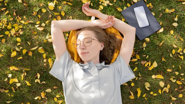 Woman relaxing on grass with yellow leaves, eyes closed, arms stretched back, next to tablet and phone.