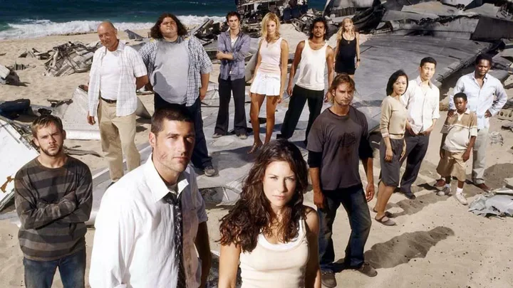Group of people stand on a beach with airplane wreckage in the background, reminiscent of a survival scenario.