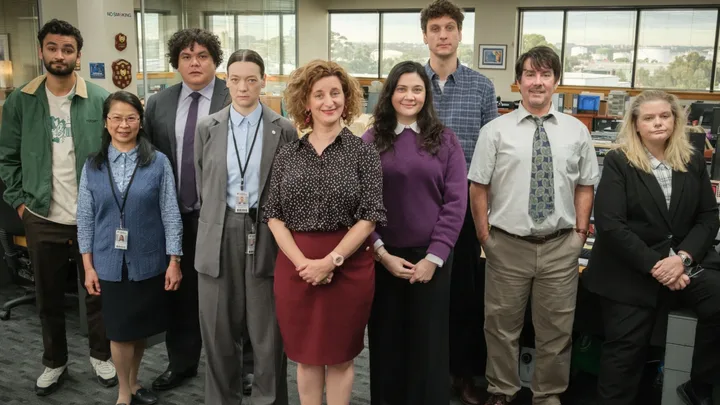 Cast of the TV show "Utopia," featuring nine people standing in an office setting.