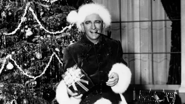 Man in Santa hat holds a gift while smiling and standing next to a Christmas tree.