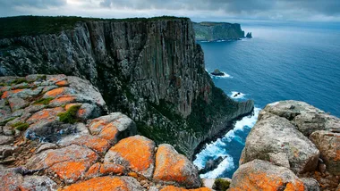 Rocky coastal cliffs covered in orange lichen overlook the ocean under a cloudy sky.