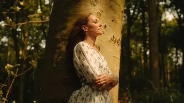 Woman in patterned dress leaning against a tree in a sunlit forest, eyes closed and arms crossed.