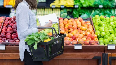 Person grocery shopping with a basket of produce, holding a shopping list, in front of apple display.