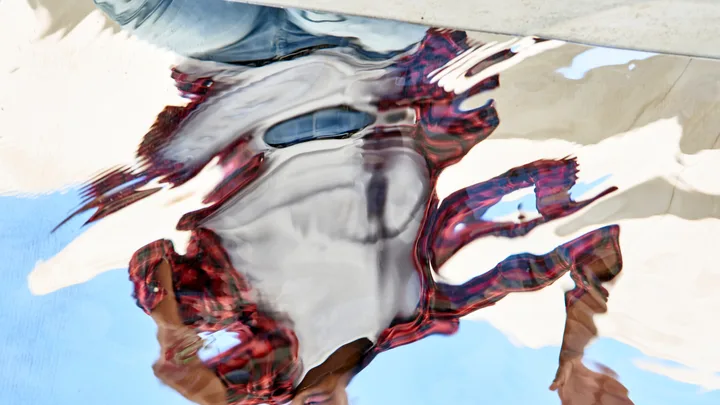 Distorted reflection of a person in water, wearing a red and black plaid shirt and white top.