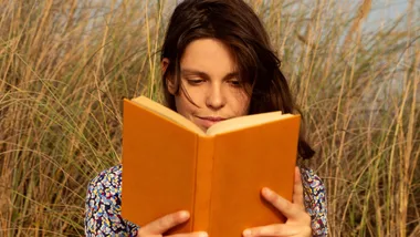 Woman reading an orange book, surrounded by tall grass outdoors.