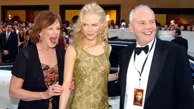 Three people dressed elegantly at a formal event, with a crowd in the background.