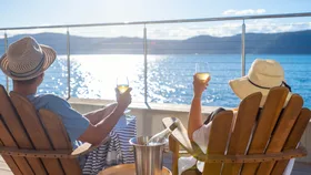 A couple in sun hats relaxes with wine on a deck overlooking a sunny lake view.