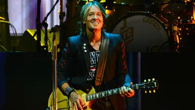 Musician performing on stage with a guitar, smiling, and wearing a patterned shirt and black jacket.
