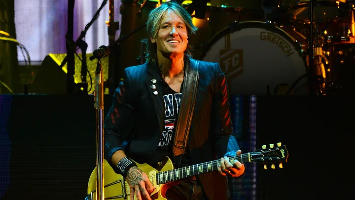Musician performing on stage with a guitar, smiling, and wearing a patterned shirt and black jacket.