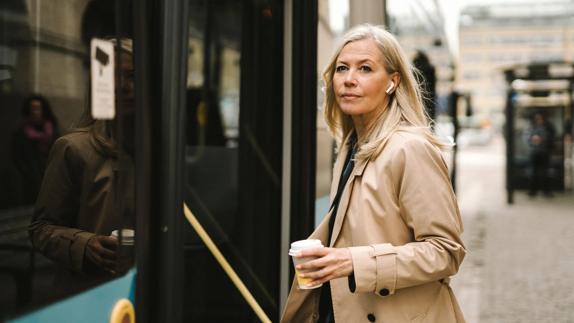 A woman with earphones boards a bus, holding a coffee cup, wearing a beige coat on an urban street.