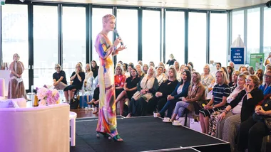 A woman speaks on stage to a seated audience in a well-lit room with large windows.