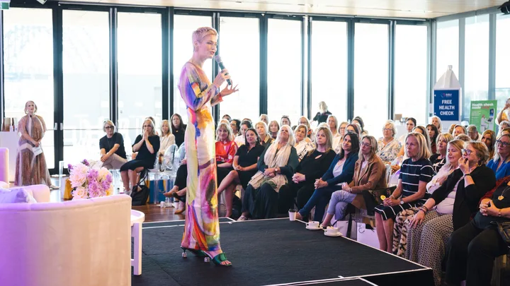 A woman speaks on stage to a seated audience in a well-lit room with large windows.