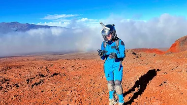 Person in blue spacesuit and helmet walking on rocky, Mars-like terrain with a clear blue sky backdrop.