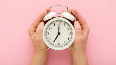 Hands holding a white alarm clock on a pink background.