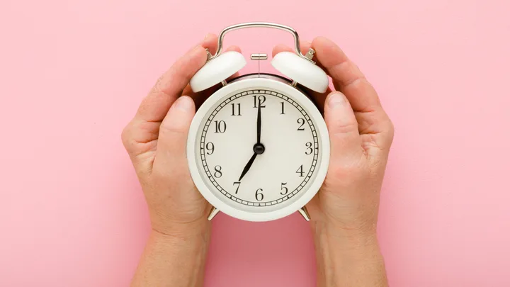 Hands holding a white alarm clock on a pink background.