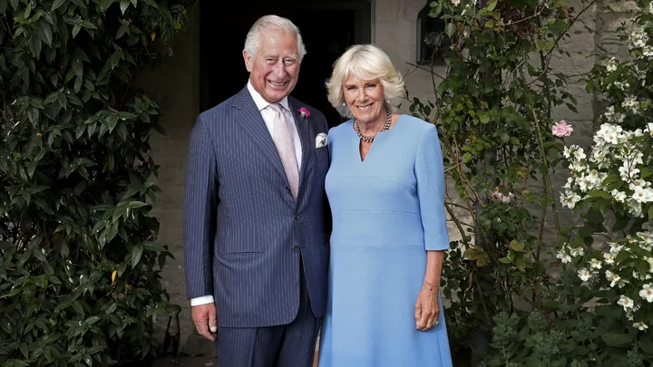 Man in pinstripe suit and woman in blue dress smiling, standing outdoors near green leafy plants and flowers.