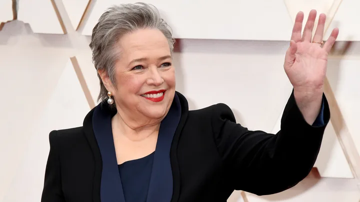 A woman in a black suit with pearl earrings smiles and waves at an event.