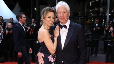 Couple on the red carpet, smiling at a formal event, with photographers in the background.