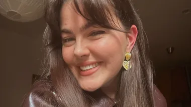 Smiling person with long hair, wearing gold heart-shaped earrings and a shiny brown jacket.