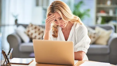 Woman looking stressed while using a laptop at home, hand on forehead, sitting in a cozy living room.