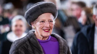 An elderly woman in a fur hat and purple outfit smiles warmly in a crowded setting.