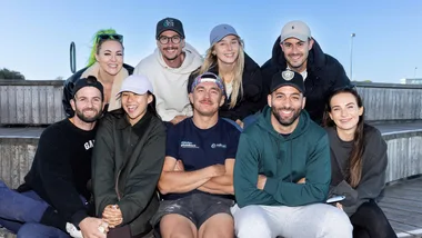 Group of nine people sitting on outdoor wooden steps, smiling and wearing casual clothing and caps on a clear day.