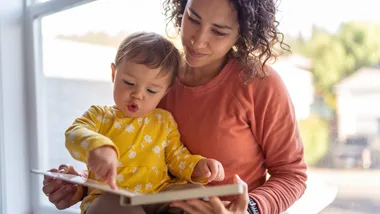 Woman and child in yellow dress reading a book by a window, with natural light and blurred background.