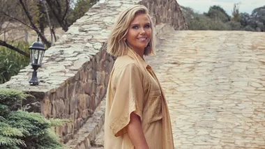 A woman in a beige outfit stands on a stone path, smiling, with greenery and a stone wall in the background.