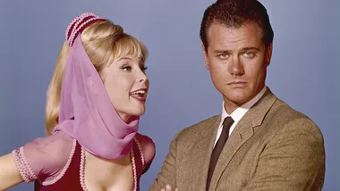 A woman in a pink outfit and veil talks to a serious man in a brown suit against a light blue background.