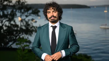 Man in a teal suit with a black tie stands in front of a scenic waterfront background with boats.