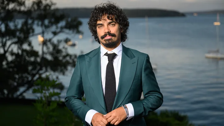 Man in a teal suit with a black tie stands in front of a scenic waterfront background with boats.